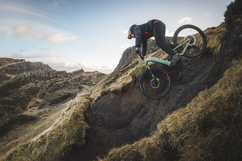 Danny MacAskill rast einen steilen und steinigen Trail herab. Die Reifen haben kaum Bodenkontakt, Im Hintergrund ist hügelige Felslandschaft.